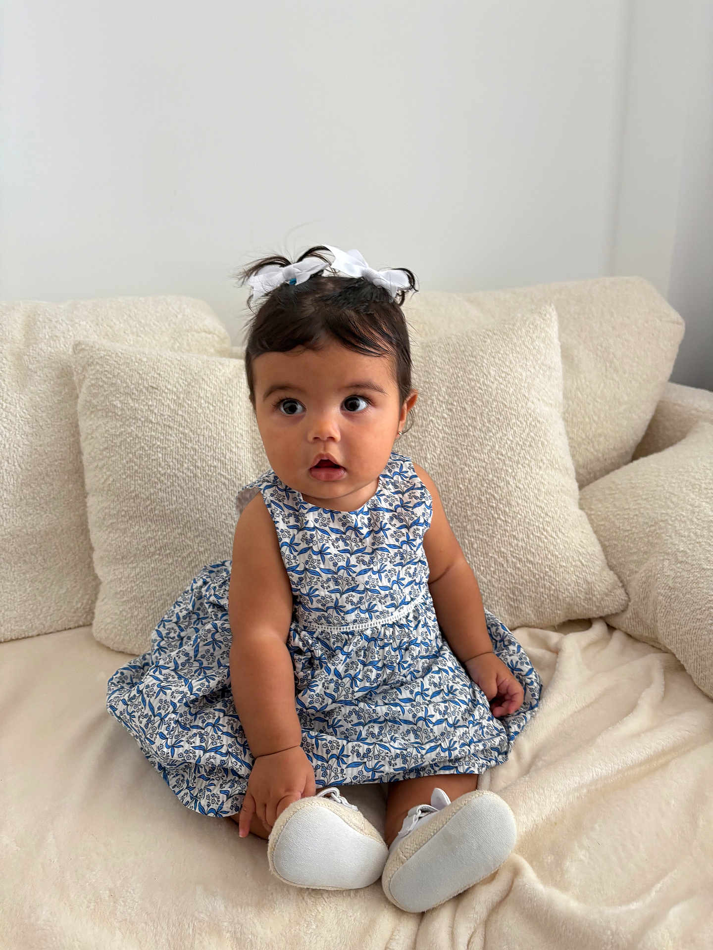 Alba a rejoint le concours — aidez-le/la à gagner de superbes lots ! toddler, child, girl, dress, blue_and_white, floral_pattern, white_bows, hair_accessories, white_shoes, couch, pillows, blanket, indoor, sitting, curious, wide_eyes, cute, portrait, baby, soft_lighting
