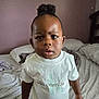 bed, bedroom, blanket, brown_skin, calvin_klein, casual_clothing, child, curly_hair, expression, face, home, indoor, looking_at_camera, person, pillows, puffed_hair, standing, toddler, white_shirt, young_child