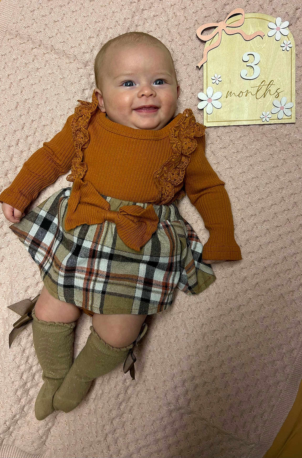 Ainsley is registered to the contest to win money with this photo: baby, infant, smiling, plaid_skirt, mustard_top, long_sleeves, knee_high_socks, bow, wooden_sign, pink_blanket, texture, flower_decorations, milestone, cute, happy, lying_down, indoors, portrait, child, person