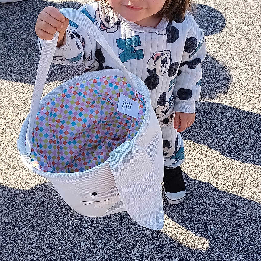 Elijio is registered to the contest to win money with this photo: basket, bunny_basket, casual_clothing, child, curious, curly_hair, cute, daylight, footwear, holding, outdoor, pavement, person, photobomb, playful, ponytail, shadow, smile, sunlight, toddler