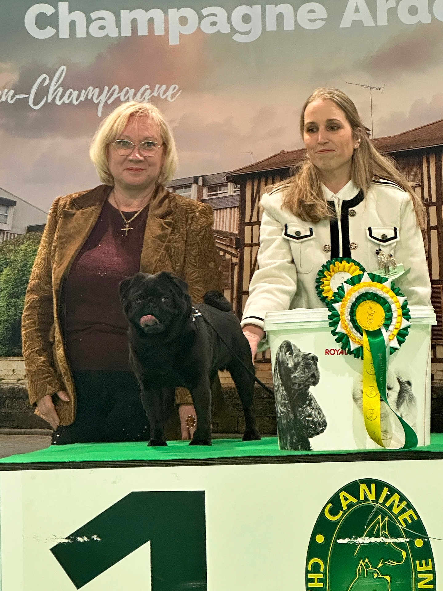 Saint Maxime a rejoint le concours — aidez-le/la à gagner de superbes lots ! dog, pug, black_dog, woman, person, award_ribbon, rosette, podium, backdrop, banner, glasses, jacket, white_jacket, brown_jacket, leash, prize_bucket, stage, portrait, smile, competition
