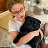 black_fur, brown_hair, child, close_up, couch, cozy, dog, eyewear, girl, glasses, harness, holding, home_interior, hug, living_room, pet, plant, portrait, pug, white_tshirt