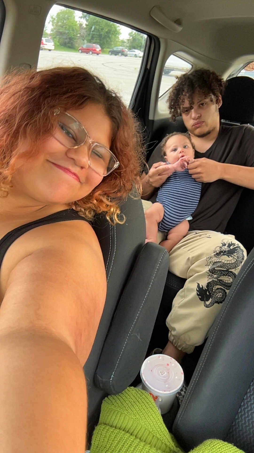 Grayson is registered to the contest to win money with this photo: car_interior, selfie, young_woman, young_man, baby, holding_baby, glasses, curly_hair, striped_onesie, parking_lot, drink_cup, green_sweater, smiling, casual_clothing, seat, window, outdoor_view, people, family_moment, portrait