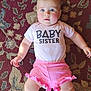 baby, infant, child, pink_shorts, pink_onesie, baby_sister_text, carpet, patterned_rug, laying_down, curious_expression, chubby_legs, blue_eyes, short_hair, indoor, cute, adorable, skin, human, person, portrait