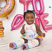 Junei is registered to the contest to win money with this photo: baby, child, smiling, curly_hair, balloons, donuts, birthday, celebration, colorful, sitting, outfit, drool, cute, decorations, happy, legs, feet, white_background, party, girl