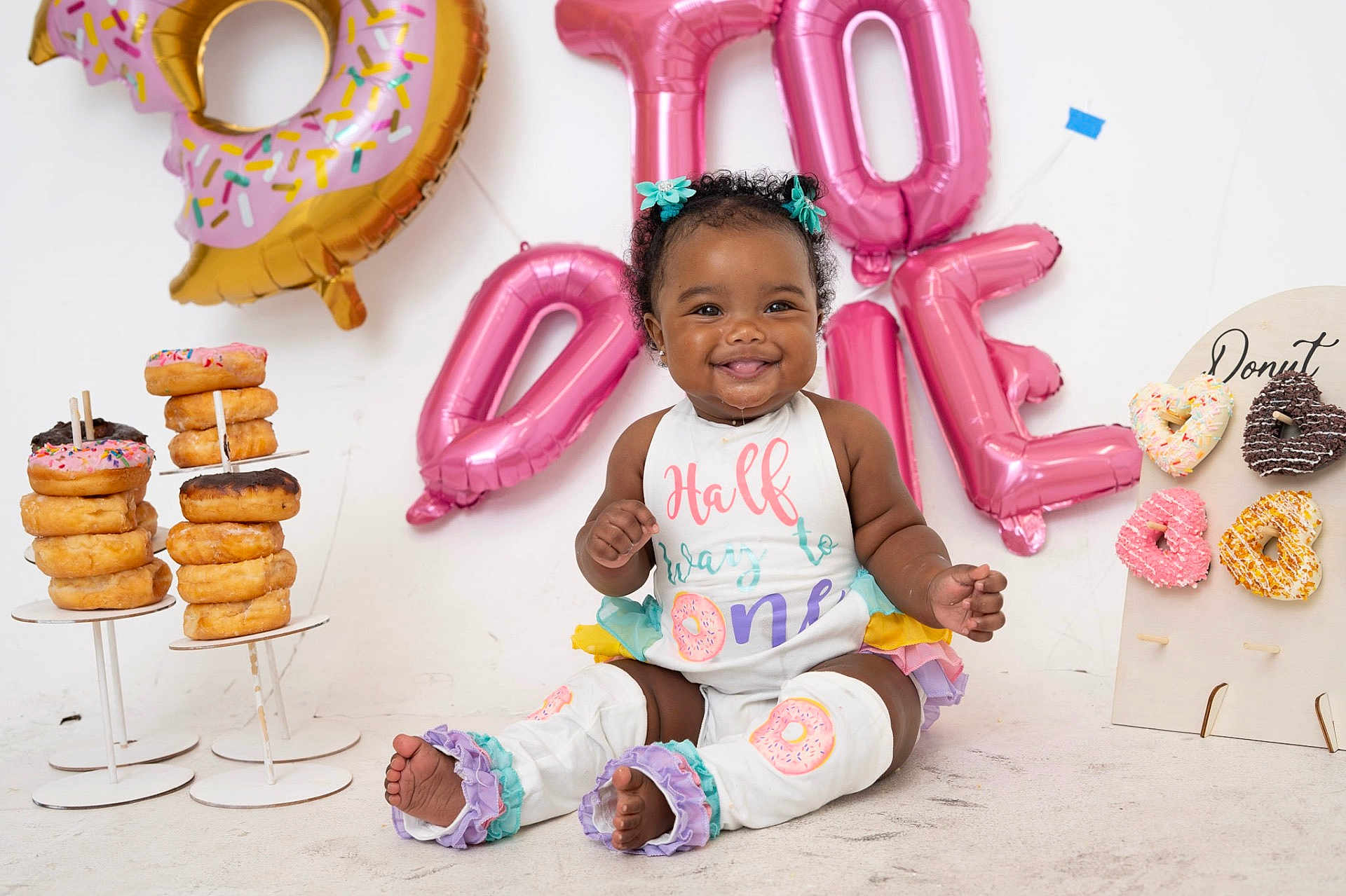 Junei is registered to the contest to win money with this photo: baby, child, smiling, curly_hair, balloons, donuts, birthday, celebration, colorful, sitting, outfit, drool, cute, decorations, happy, legs, feet, white_background, party, girl