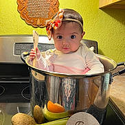 Milena is registered to the contest to win money with this photo: baby, infant, kitchen, pot, vegetables, carrot, corn, potato, celery, wooden_spatula, headband, bow, stove, countertop, sign, five_months, prayer_plaque, green_wall, cute, child