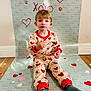 child, toddler, boy, pajamas, heart_pattern, valentine_backdrop, sitting, socks, hardwood_floor, paper_hearts, love_text, kiss_text, backdrop, wall, portrait, indoor, cute, expression_neutral, flooring, studio_setup