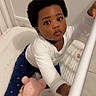 toddler, child, baby, crib, toy, stuffed_animal, curly_hair, white_shirt, blue_pants, pink_shoes, floor, indoor, cute, looking_up, hand_rail, playful, infant, person, home, young_child