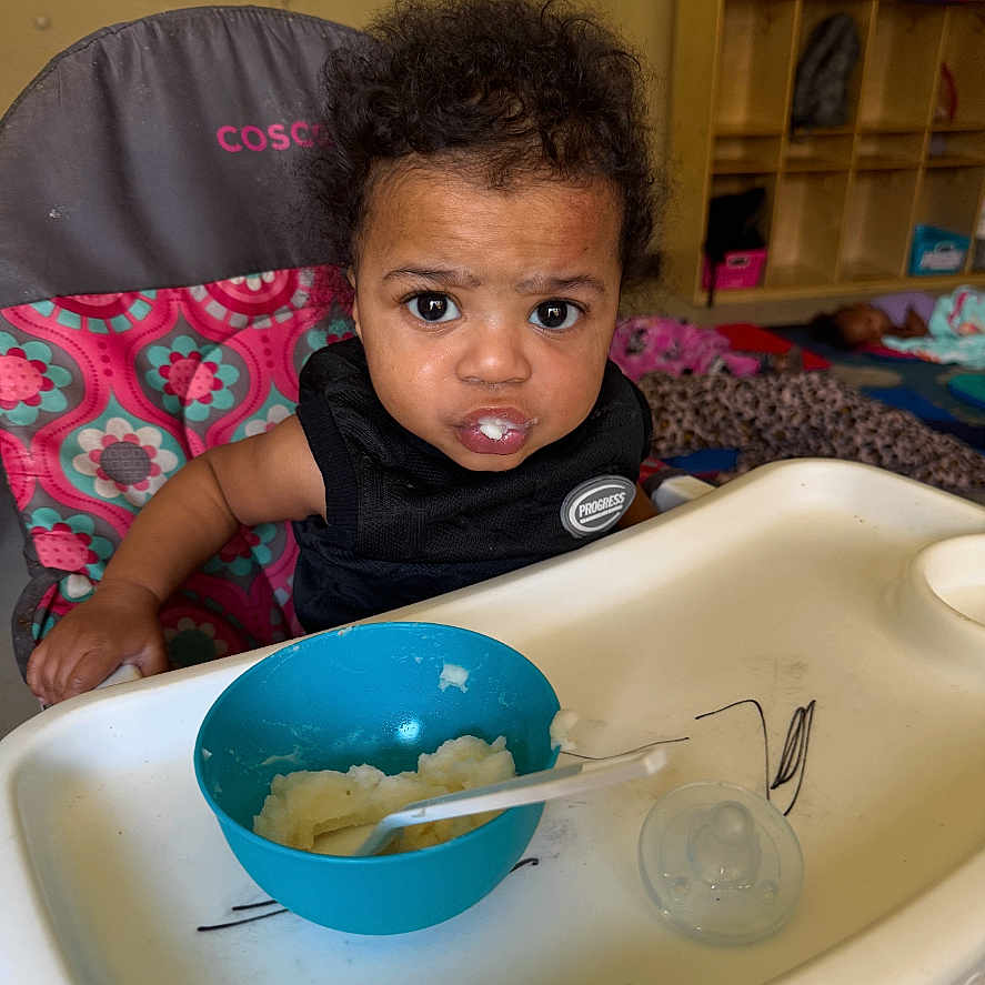 Dkhaza is registered to the contest to win money with this photo: background, black_clothing, bowl, child, colorful, curly_hair, daycare, expression, face, feeding, food, furniture, high_chair, indoor, pacifier, person, room, sitting, spoon, toddler