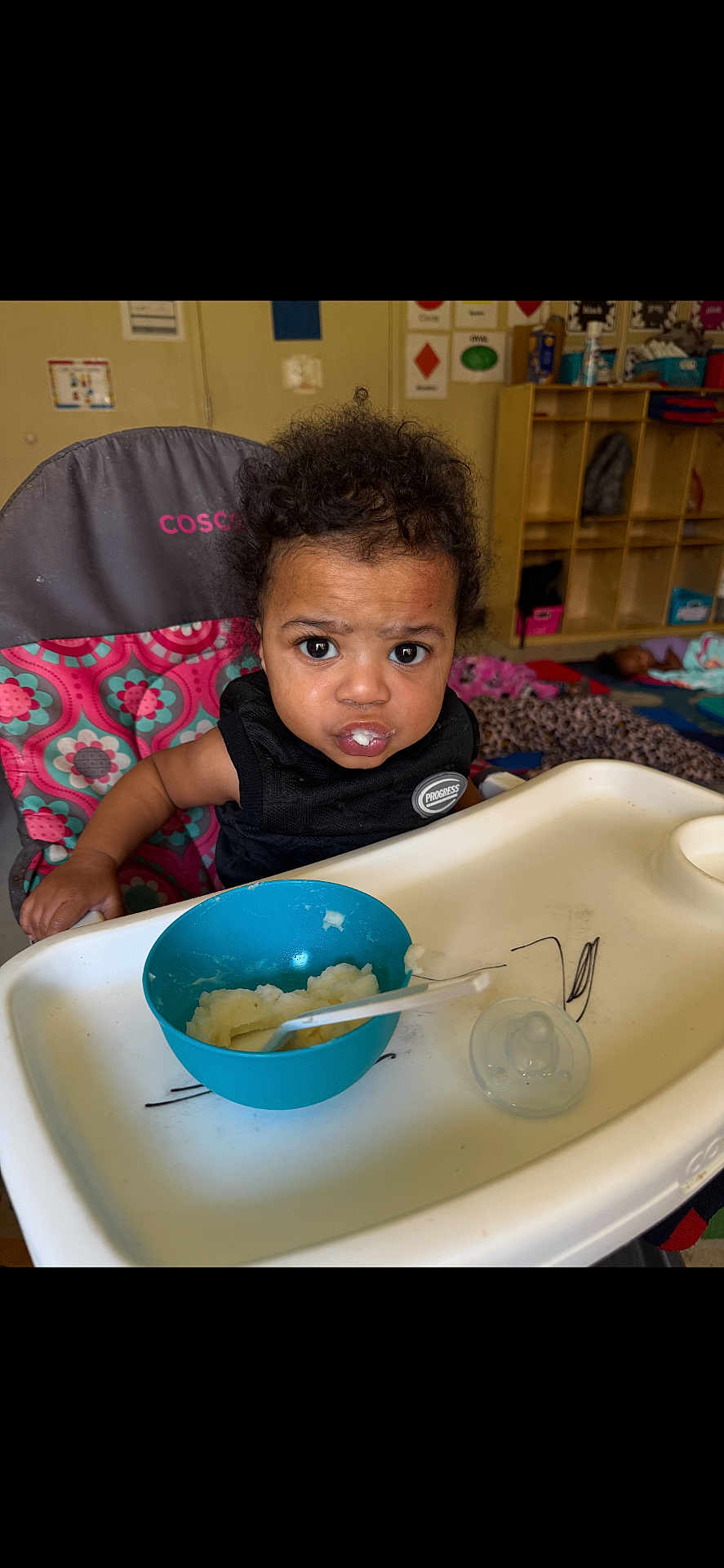 Dkhaza is registered to the contest to win money with this photo: toddler, high_chair, food, bowl, spoon, indoor, child, curly_hair, black_clothing, expression, face, feeding, pacifier, furniture, room, sitting, colorful, daycare, background, person