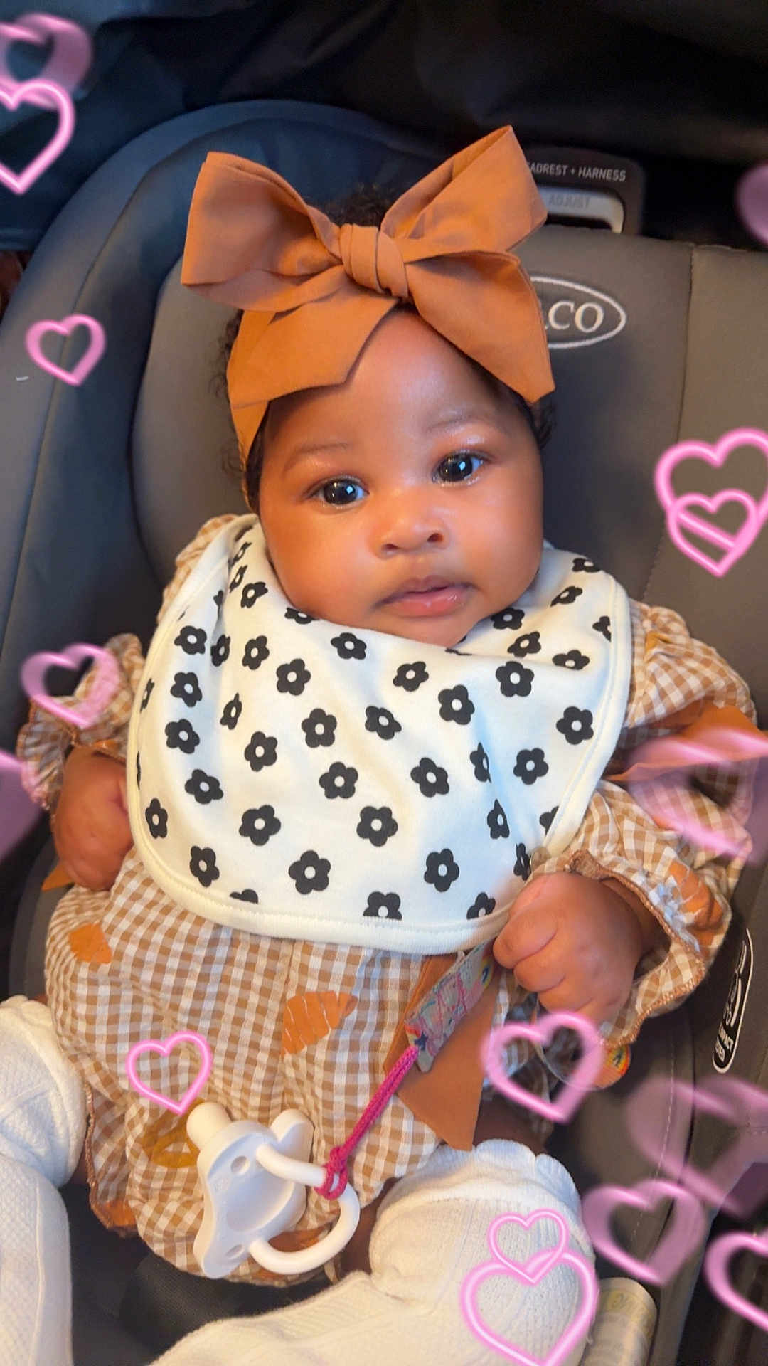 Keilani is registered to the contest to win money with this photo: baby, child, car_seat, headband, bow, bib, pacifier, clothing, patterned_bib, patterned_clothing, pink_hearts, infant, cute, portrait, indoor, person, sitting, closeup, adorable, soft_light