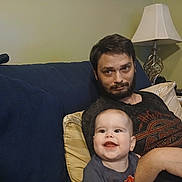 Roselynn is registered to the contest to win money with this photo: man, baby, couch, lamp, pillow, living_room, beard, smile, happy, indoor, casual, relaxing, person, furniture, home, portrait, seated, clothing, adult, child