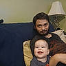 man, baby, couch, lamp, pillow, living_room, beard, smile, happy, indoor, casual, relaxing, person, furniture, home, portrait, seated, clothing, adult, child