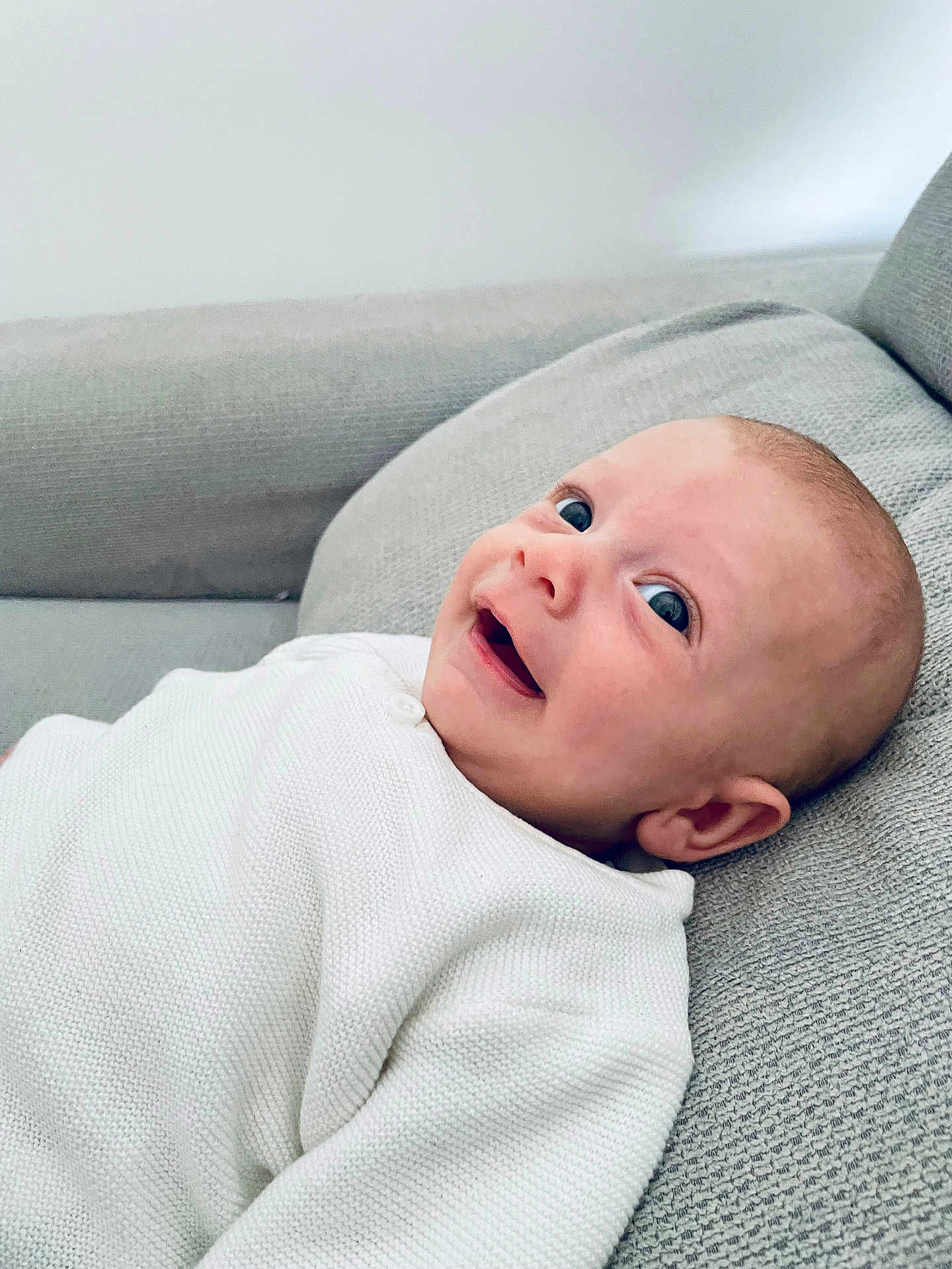 Noam a rejoint le concours — aidez-le/la à gagner de superbes lots ! baby, infant, smile, happy, face, white_clothing, knitwear, couch, gray_fabric, indoors, person, head, skin, cute, child, lying_down, closeup, young, cozy, comfort