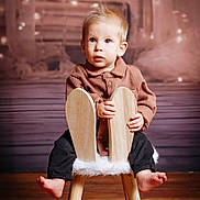 Maëlio a rejoint le concours — aidez-le/la à gagner de superbes lots ! toddler, child, barefoot, wooden_chair, fluffy, brown_jacket, black_pants, indoor, portrait, curious_expression, blond_hair, blue_eyes, soft_lighting, wooden_floor, decorative_background, seated, cute, small_hands, young_child, cozy