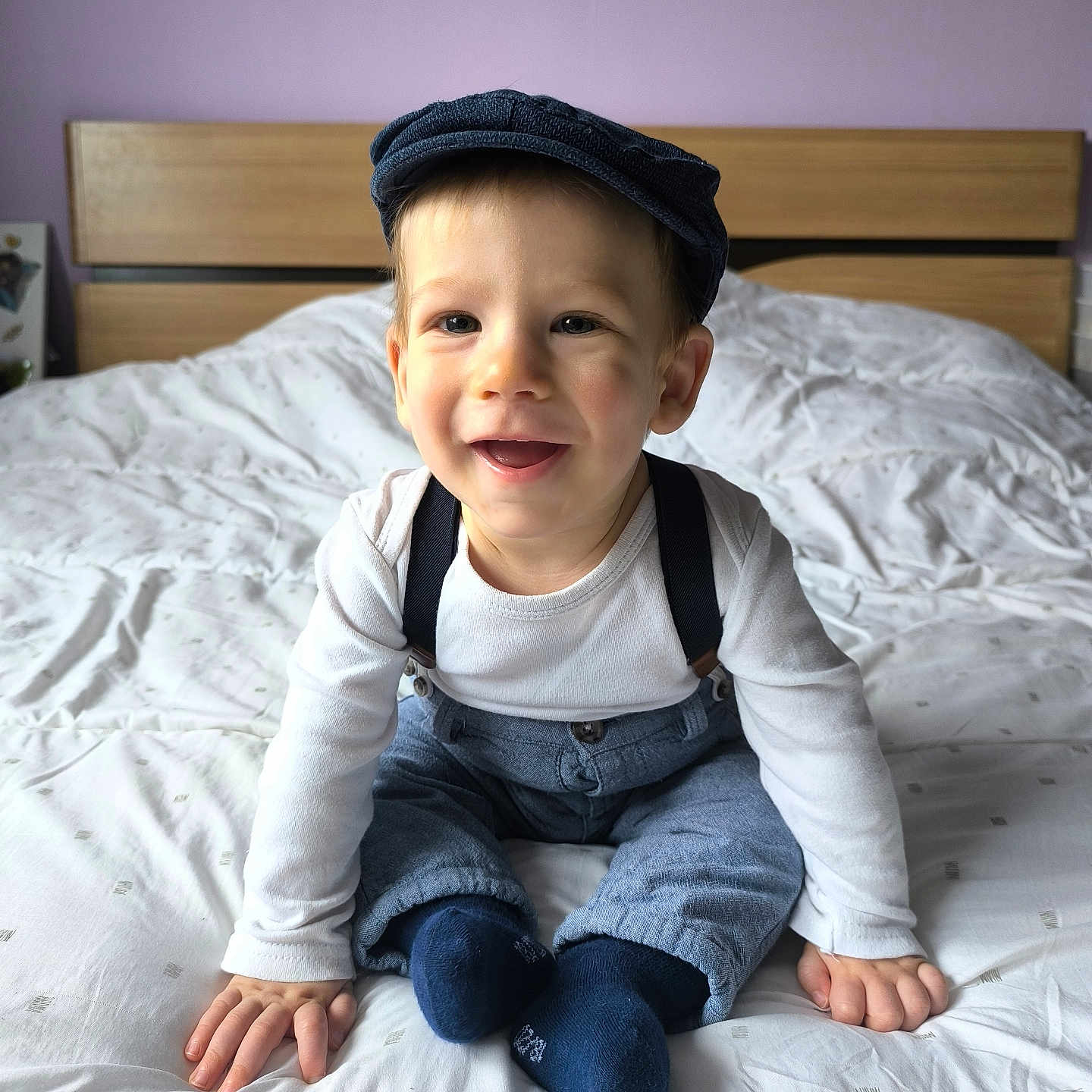 Maëlio participe au concours pour gagner de l'argent avec cette photo : baby, bed, blanket, blue_pants, blue_socks, cap, child, cute, face, happy, headwear, home, indoor, person, portrait, sitting, smiling, suspenders, toddler, white_shirt