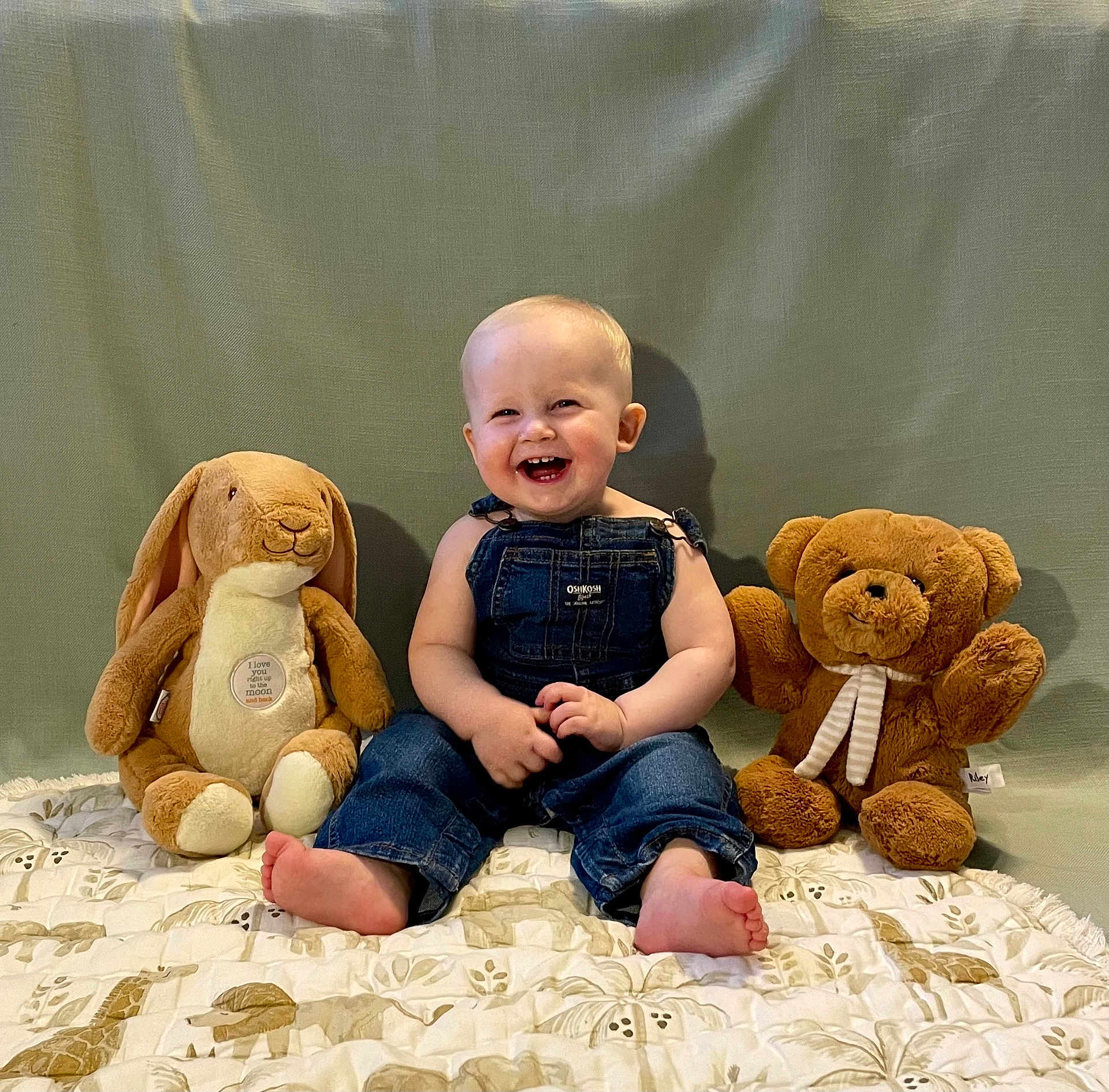 Thomas is registered to the contest to win money with this photo: baby, child, denim_overalls, smiling, happy, sitting, plush_bunny, teddy_bear, quilt, blanket, indoor, portrait, cute, barefoot, toy, soft_toy, green_background, infant, joyful, cuddly