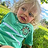 Hudson is registered to the contest to win money with this photo: child, toddler, curly_hair, green_shirt, outdoor, grass, sunny, blue_sky, tree, nature, portrait, casual_clothing, young_child, daylight, face, person, expression, hand, sitting, relaxed
