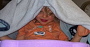 Hudson is registered to the contest to win money with this photo: child, towel, high_chair, orange_tshirt, beige_shorts, indoor, smiling, playful, face, person, furniture, ceiling_fan, home, curly_hair, table, seat, blanket, cozy, cute, kid