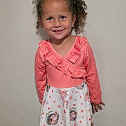 Ariela is registered to the contest to win money with this photo: child, girl, smile, curly_hair, dress, pink, white, floral_pattern, deer_pattern, long_sleeve, standing, earrings, happy, portrait, indoor, fashion, cute, person, clothing, wall