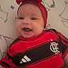 Kyara is registered to the contest to win money with this photo: baby, child, infant, smiling, happy, portrait, close_up, red_bow, headband, sports_jersey, football_jersey, red_clothing, black_clothing, adidas_logo, team_logo, sitting, cushion, indoor, bedroom, patterned_background