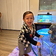Daniel is registered to the contest to win money with this photo: toddler, child, laughing, indoor, glass_table, purple_rug, checkered_jacket, boots, fish_tank, living_room, smiling, wooden_floor, window_blinds, person, home_decor, casual_clothing, happy, young_child, furniture, playful