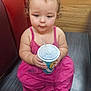 Willow is registered to the contest to win money with this photo: toddler, child, pink_dress, cup, straw, sitting, floor, indoor, curly_hair, cross_legged, red_bench, wooden_wall, barefoot, cute, drinking, young_child, casual, portrait, baby, contemplative