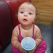 Willow is registered to the contest to win money with this photo: toddler, child, cup, straw, pink_clothing, curly_hair, blue_eyes, floor, wooden_wall, red_leather_seat, indoor, expression, looking_up, holding, sitting, curious, rosy_cheeks, cute, innocent, casual