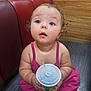 toddler, child, cup, straw, pink_clothing, curly_hair, blue_eyes, floor, wooden_wall, red_leather_seat, indoor, expression, looking_up, holding, sitting, curious, rosy_cheeks, cute, innocent, casual