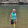 child, boy, water, life_vest, swim_trunks, smile, standing, outdoor, river, rocks, nature, summer, fun, recreation, water_safety, happy, portrait, adventure, shallow_water, playful