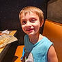 child, boy, smile, missing_tooth, restaurant, booth, table, food, fries, blue_shirt, dinosaur_graphic, short_hair, casual_clothing, indoor, portrait, happy, seated, light_skin, face, person