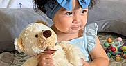 Amelia Guerrero joined the competition — help win amazing prizes! toddler, child, curly_hair, blue_bow, headband, teddy_bear, plush_toy, bed, bedding, toy, colorful, cute, indoor, person, smile, sitting, soft, cozy, playful, young_child