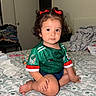 Amelia Guerrero is registered to the contest to win money with this photo: toddler, child, baby, curly_hair, red_bows, green_jersey, soccer_shirt, mexico, bed, bedspread, indoor, room, clothing, patterned_bedspread, sitting, person, cute, portrait, casual, home