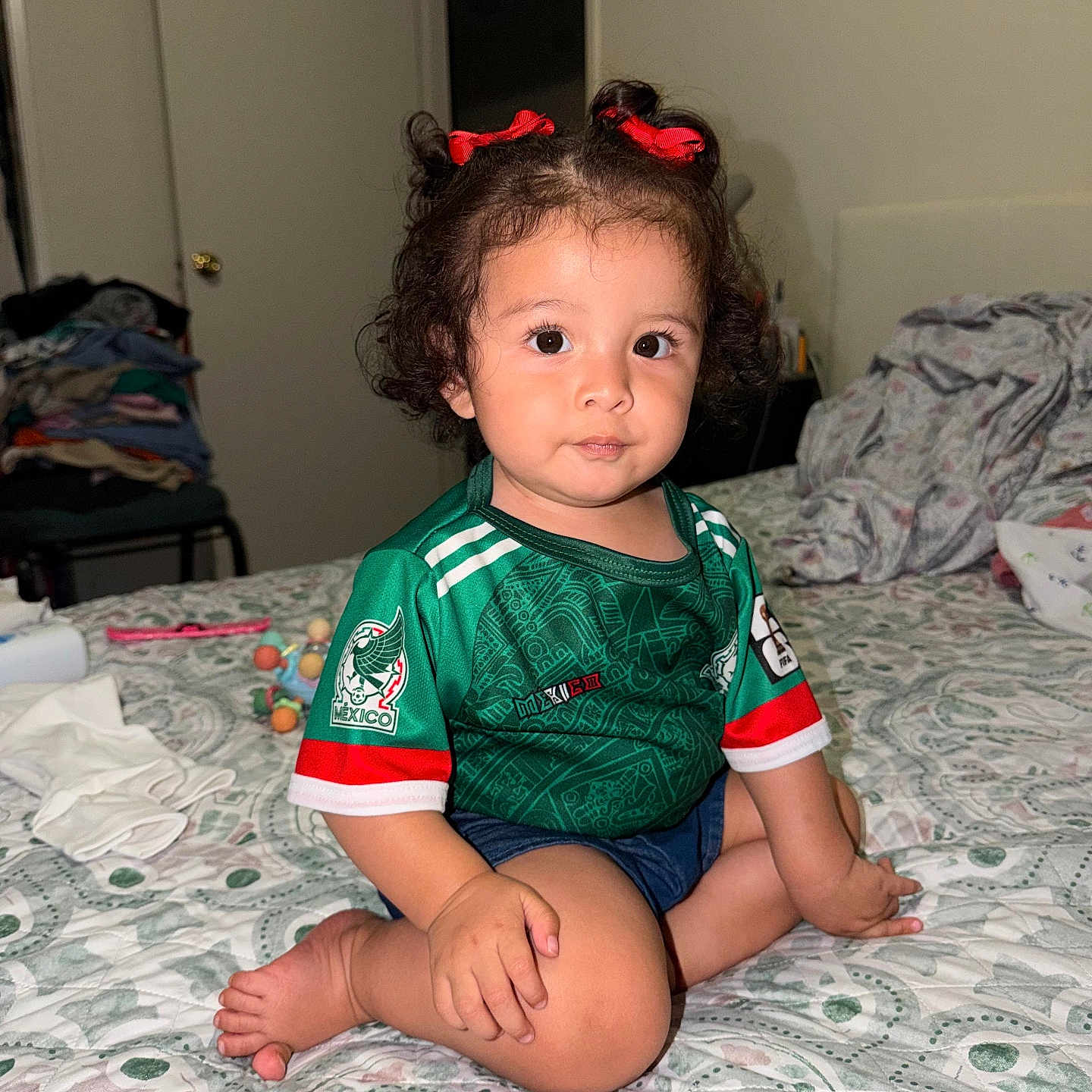 Amelia Guerrero is registered to the contest to win money with this photo: toddler, child, baby, curly_hair, red_bows, green_jersey, soccer_shirt, mexico, bed, bedspread, indoor, room, clothing, patterned_bedspread, sitting, person, cute, portrait, casual, home