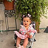 toddler, child, girl, pink_dress, sandals, hair_bows, chair, potted_plants, greenery, outdoor, curious, sitting, footwear, baby, cute, small_chair, floor, plant_stand, portrait, young_child