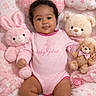 Amira is registered to the contest to win money with this photo: baby, infant, pink_onesie, teddy_bear, plush_bunny, stuffed_animals, heart_pillow, cloud_pillow, pink_theme, soft_toys, cute, smiling, child, laying_down, indoors, patterned_blanket, adorable, baby_girl, cozy, comfort