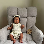 Amira joined the competition — help win amazing prizes! baby, infant, child, armchair, plush_furniture, milestone_sign, headband, cream_outfit, smiling, sitting, indoors, cozy, cute, young_child, furniture, portrait, happy, three_months, wooden_sign, texture