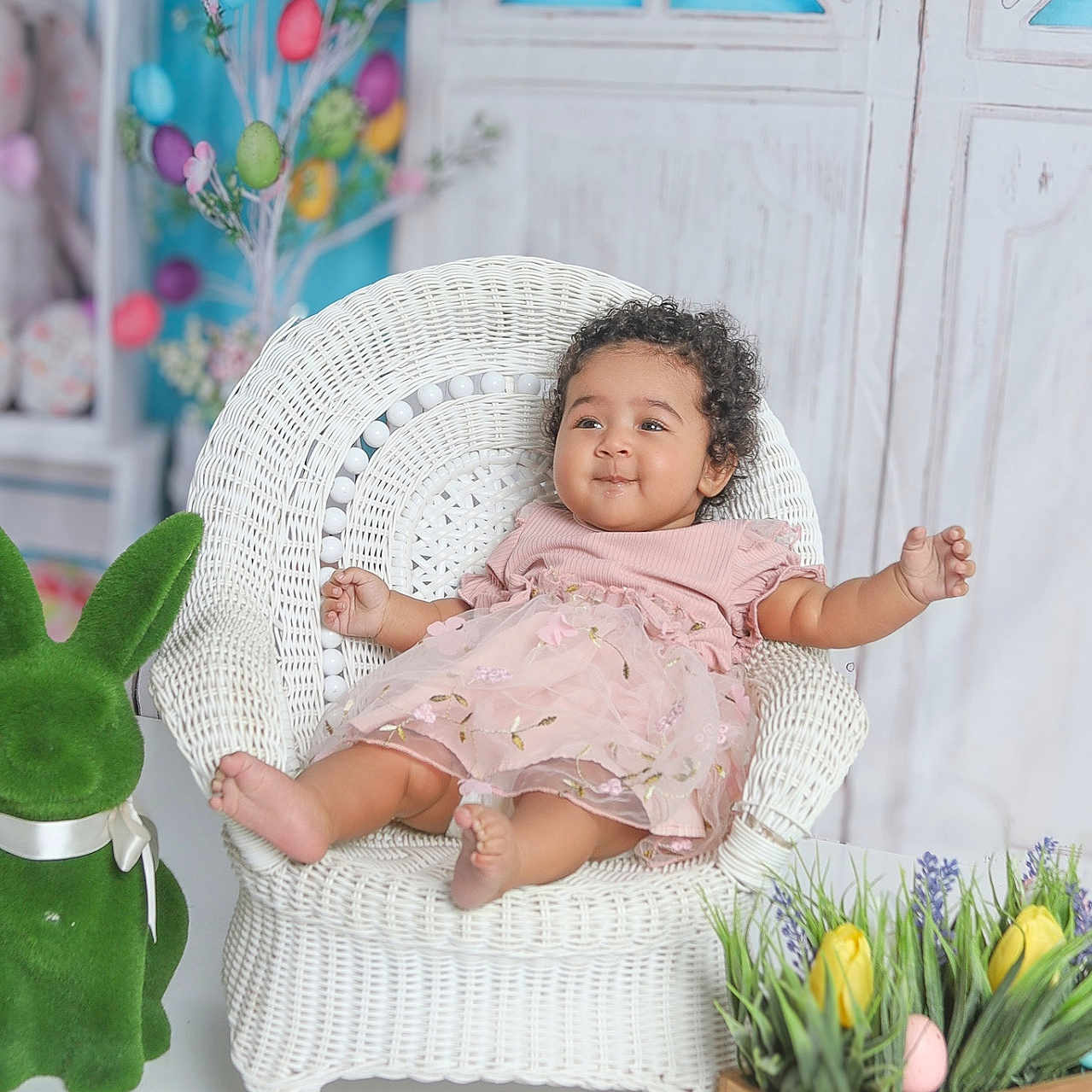 Amira joined the competition — help win amazing prizes! baby, child, wicker_chair, pink_dress, flower_embroidery, easter_eggs, green_bunny, wooden_planter, yellow_tulips, spring, decorations, indoor, smiling, curly_hair, white_background, playful, cute, festive, plants, seated