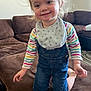 Faith is registered to the contest to win money with this photo: toddler, child, baby, overalls, denim, bib, striped_shirt, smile, kneeling, couch, living_room, brown_sofa, hair, rosy_cheeks, playful, indoor, face, eyes, hands, clothing