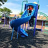 child, playground, slide, sliding, outdoor, sunny, blue_slide, orange_shirt, playground_equipment, climbing_bars, swings, trees, sky, clouds, brick_building, rubber_mulch, recreation, summer, portrait, fun
