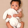 baby, child, stuffed_animal, plush_bunny, curly_hair, sitting, beige_background, soft_toy, cute, infant, portrait, small_feet, holding, comfort, adorable, expression, indoor, young_child, casual_clothing, curious