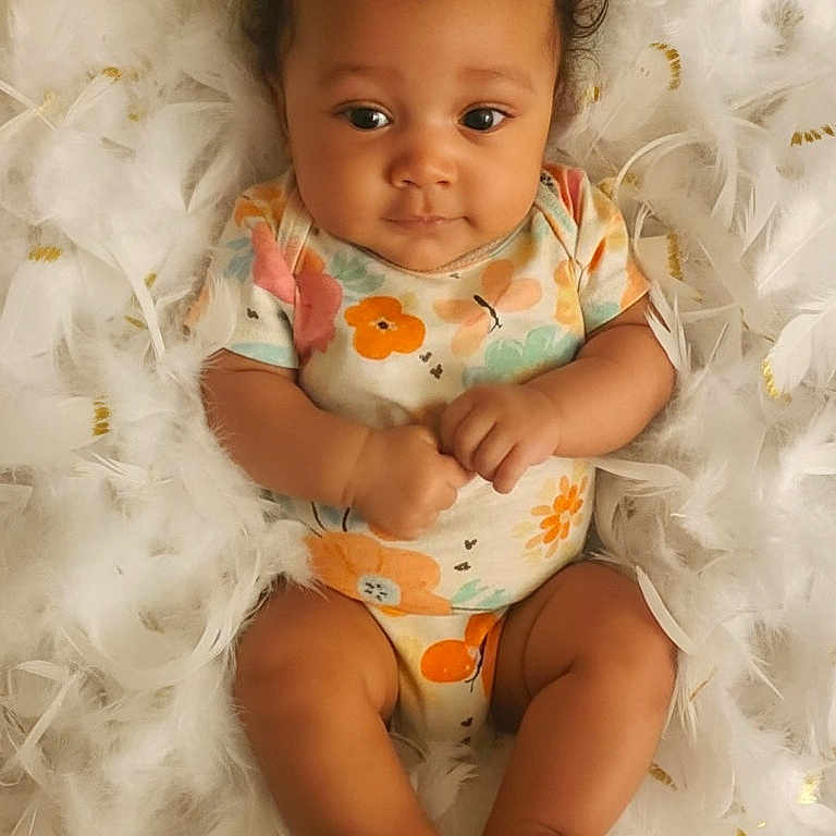 Kaiyah joined the competition — help win amazing prizes! baby, infant, child, feathers, fluffy, onesie, colorful_clothing, cute, lying_down, portrait, soft_texture, skin, small_feet, hands, curly_hair, indoors, adorable, peaceful, face, expression