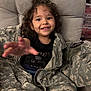 camouflage, casual, child, clothing, cozy, curly_hair, cute, fabric, girl, happy, indoors, jacket, military_jacket, person, playful, portrait, reaching_hand, seat, smile, young