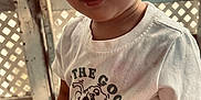 Weston is registered to the contest to win money with this photo: child, toddler, face, white_tshirt, graphic_tshirt, dinosaur, skateboard, text_on_clothing, curious_expression, closeup, wooden_background, lattice, natural_light, casual_clothing, person, young_child, indoor, cute, portrait, smile