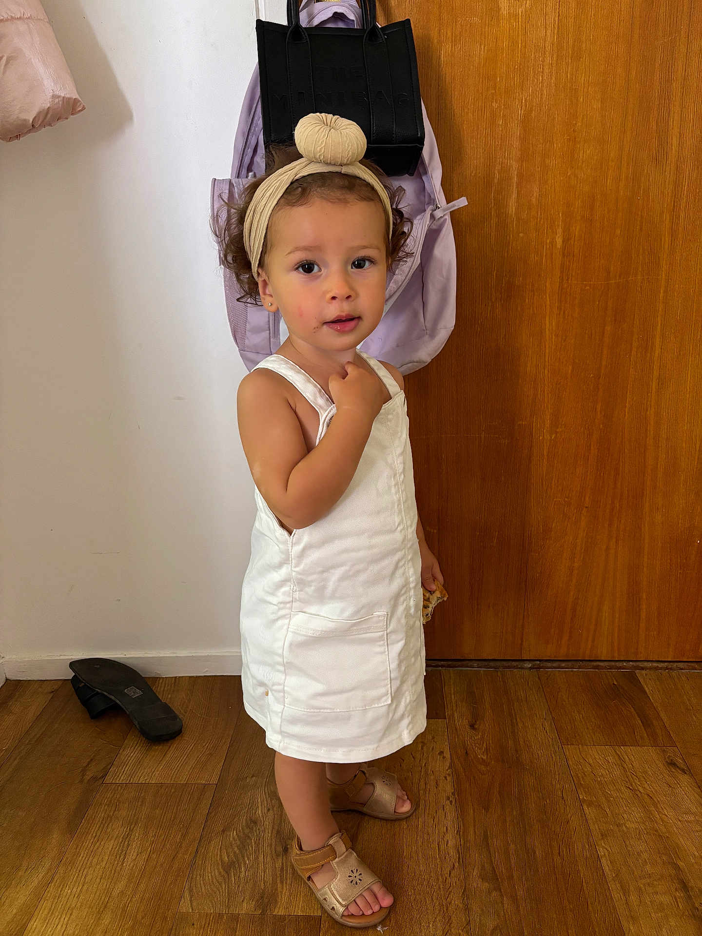 Alma a rejoint le concours — aidez-le/la à gagner de superbes lots ! toddler, child, white_dress, headband, curly_hair, sandal, wooden_floor, backpack, black_bag, door, indoor, standing, looking, hand_gesture, floor, footwear, person, young_child, casual_clothing, light_skin