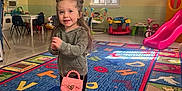 Maggie joined the competition — help win amazing prizes! alphabet_rug, chair, child, colorful_rug, cowboy_boots, floor, girl, hair_buns, hoodie, indoor, pink_purse, play_area, playroom, slide, smiling, table, toddler, toys, window, young_child