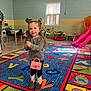 alphabet_rug, chair, child, colorful_rug, cowboy_boots, floor, girl, hair_buns, hoodie, indoor, pink_purse, play_area, playroom, slide, smiling, table, toddler, toys, window, young_child