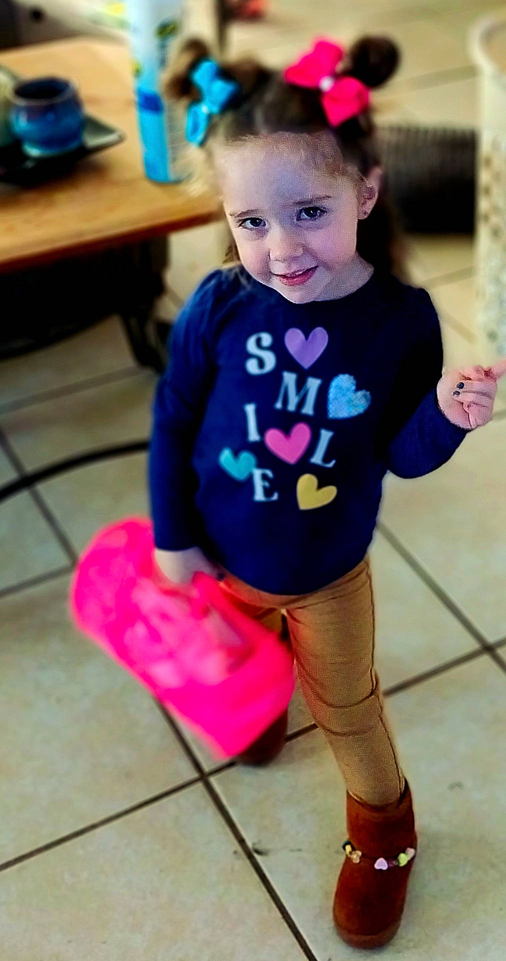 brown_boots, casual_clothing, child, colorful_bows, cute, fashion, girl, happy, hearts, indoor, navy_shirt, person, pink_bag, playful, portrait, smiling, standing, tan_pants, tiled_floor, young_child