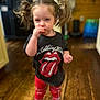 casual_clothing, child, cute, eating, expression, hair_ties, home, indoor, person, pigtails, plaid, portrait, red_pants, rolling_stones_tshirt, small_child, snack, standing, toddler, wooden_floor, young_child